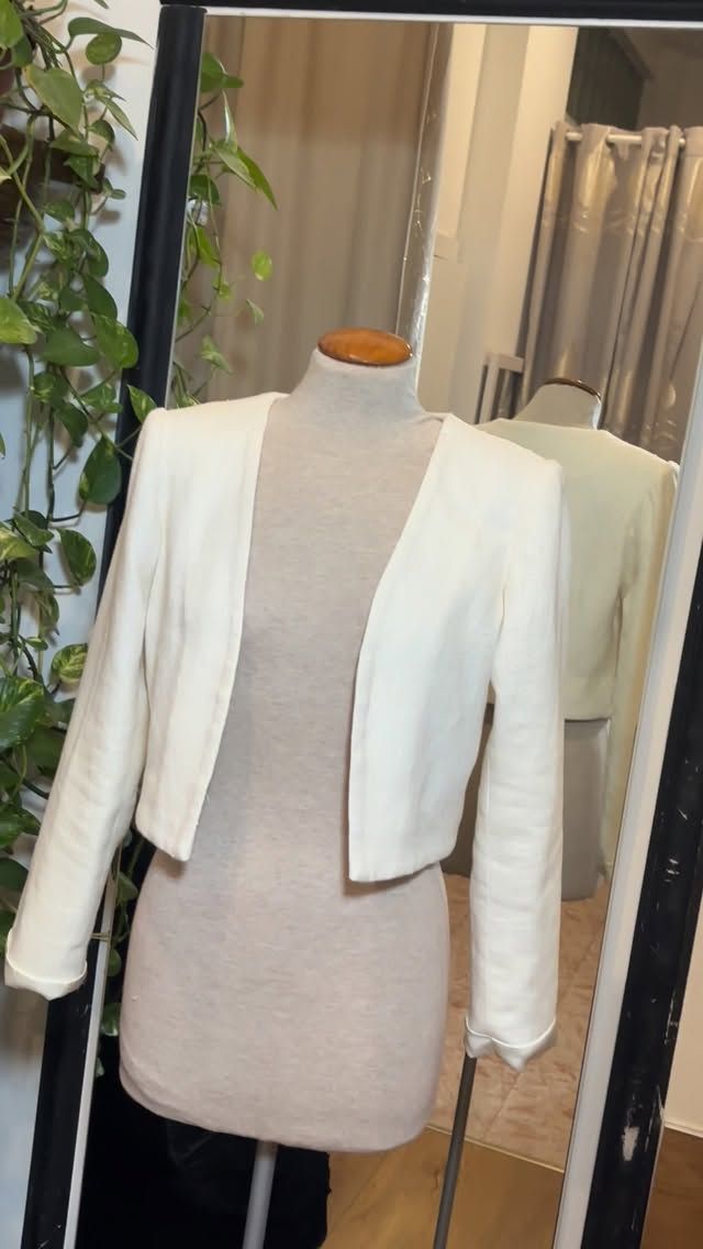 Blazer blanco corto sobre un maniquí frente a un espejo, con vegetación a la izquierda.