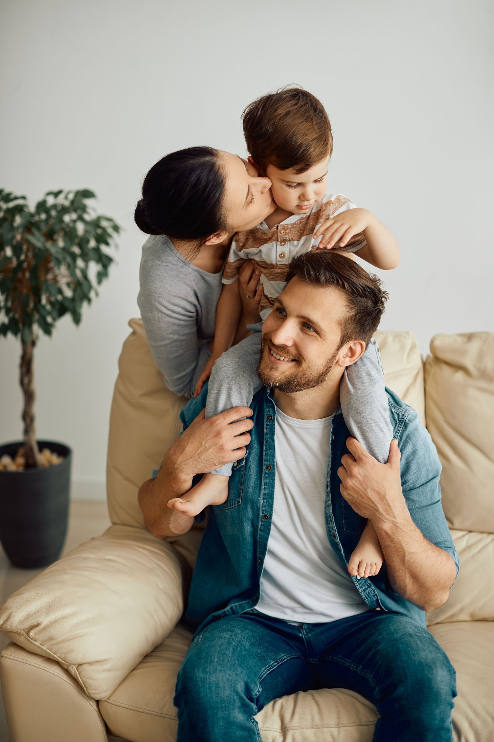 Une famille souriante sur un canapé beige, un enfant assis sur les épaules d'un homme tandis qu'une femme les enlace par derrière