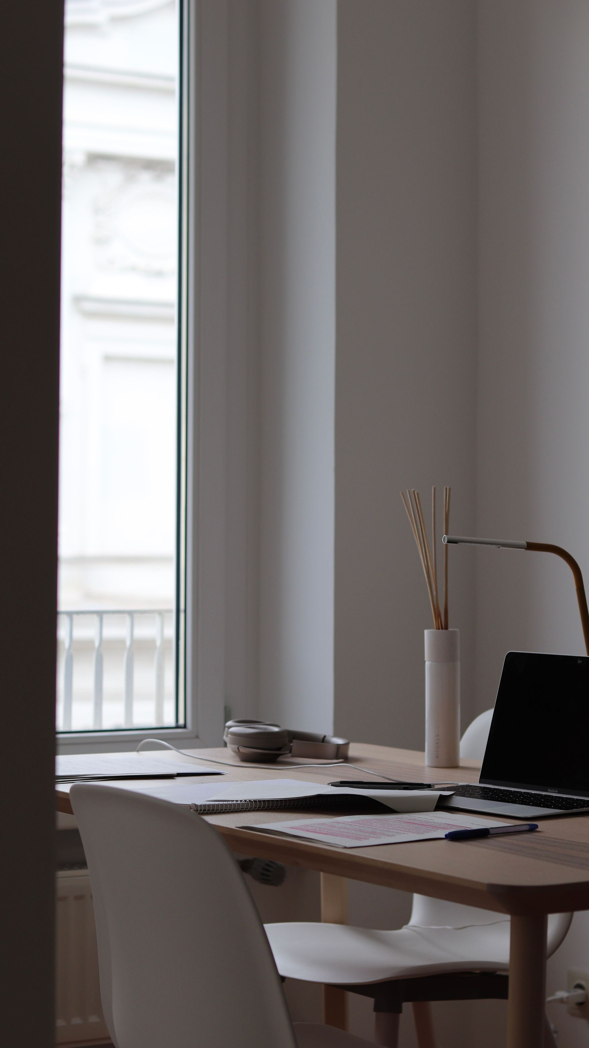 Table à manger minimaliste près d'une fenêtre avec un ordinateur portable, des papiers et un vase dans une pièce à l'éclairage tamisé