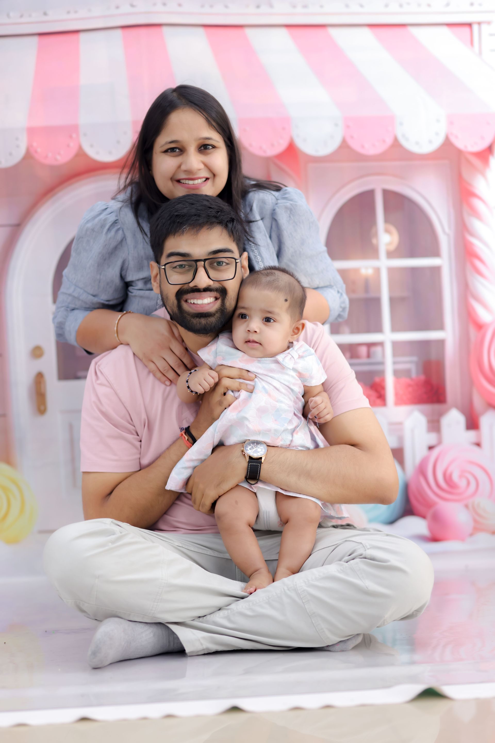 Une famille assise ensemble sur un coussin devant un fond à rayures roses et blanches, souriant à l'objectif.