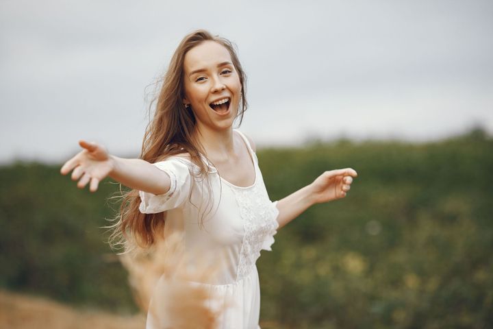 Femme souriante en robe blanche, les bras écartés, dans un champ herbeux à l'extérieur