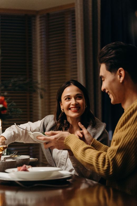 Deux personnes sourient et discutent en dînant à une table chaleureusement éclairée, avec des assiettes et des bols.
