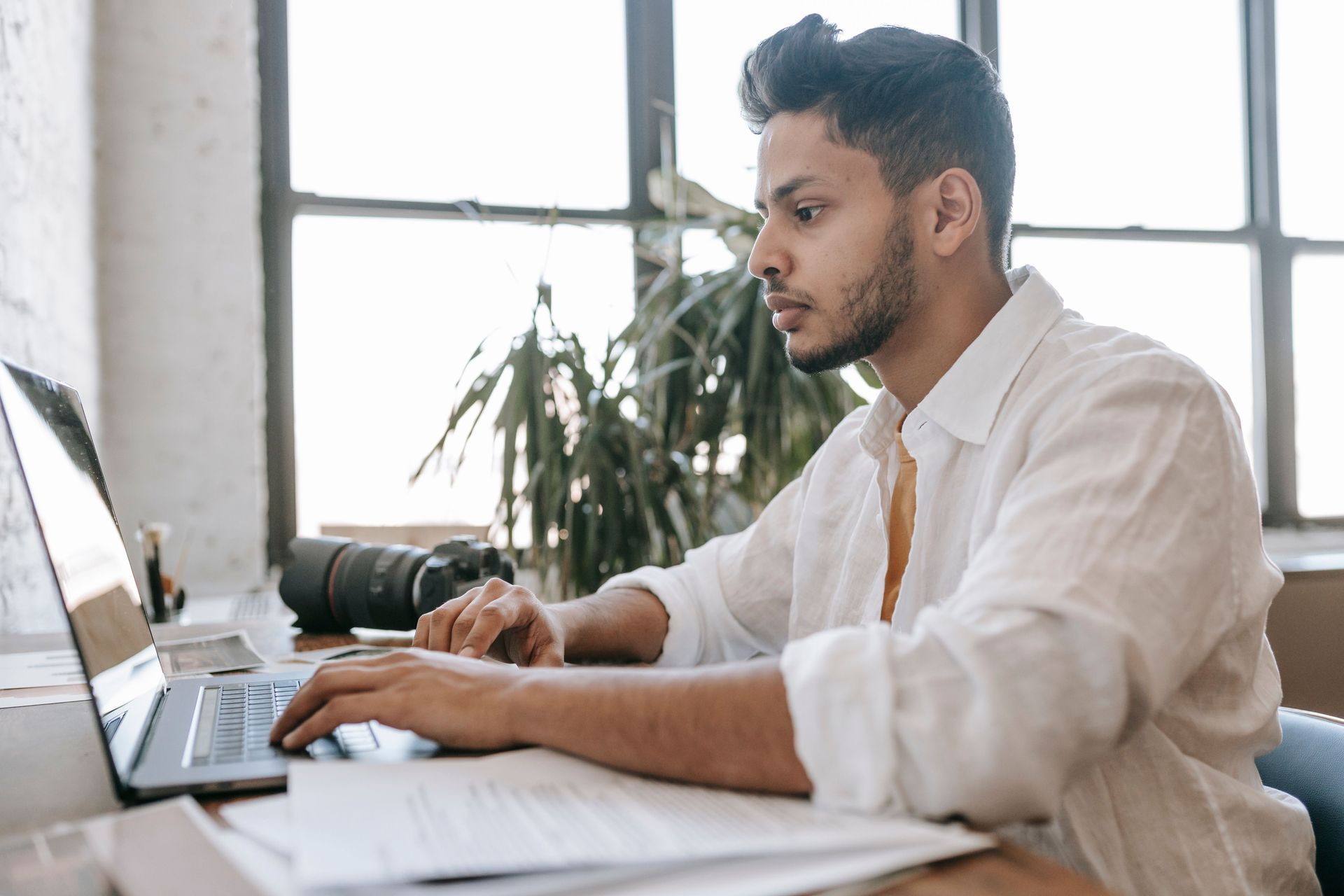 Un homme travaille sur un ordinateur portable à un bureau dans un bureau lumineux, face à de grandes fenêtres.