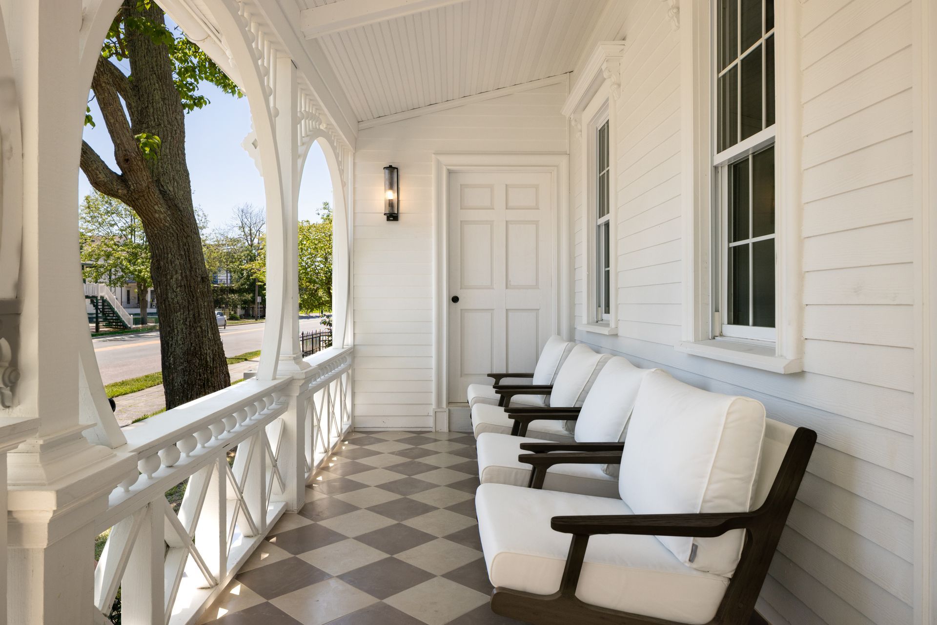 White front porch with wicker chairs, checkered floor, and a paneled door along a house exterior.