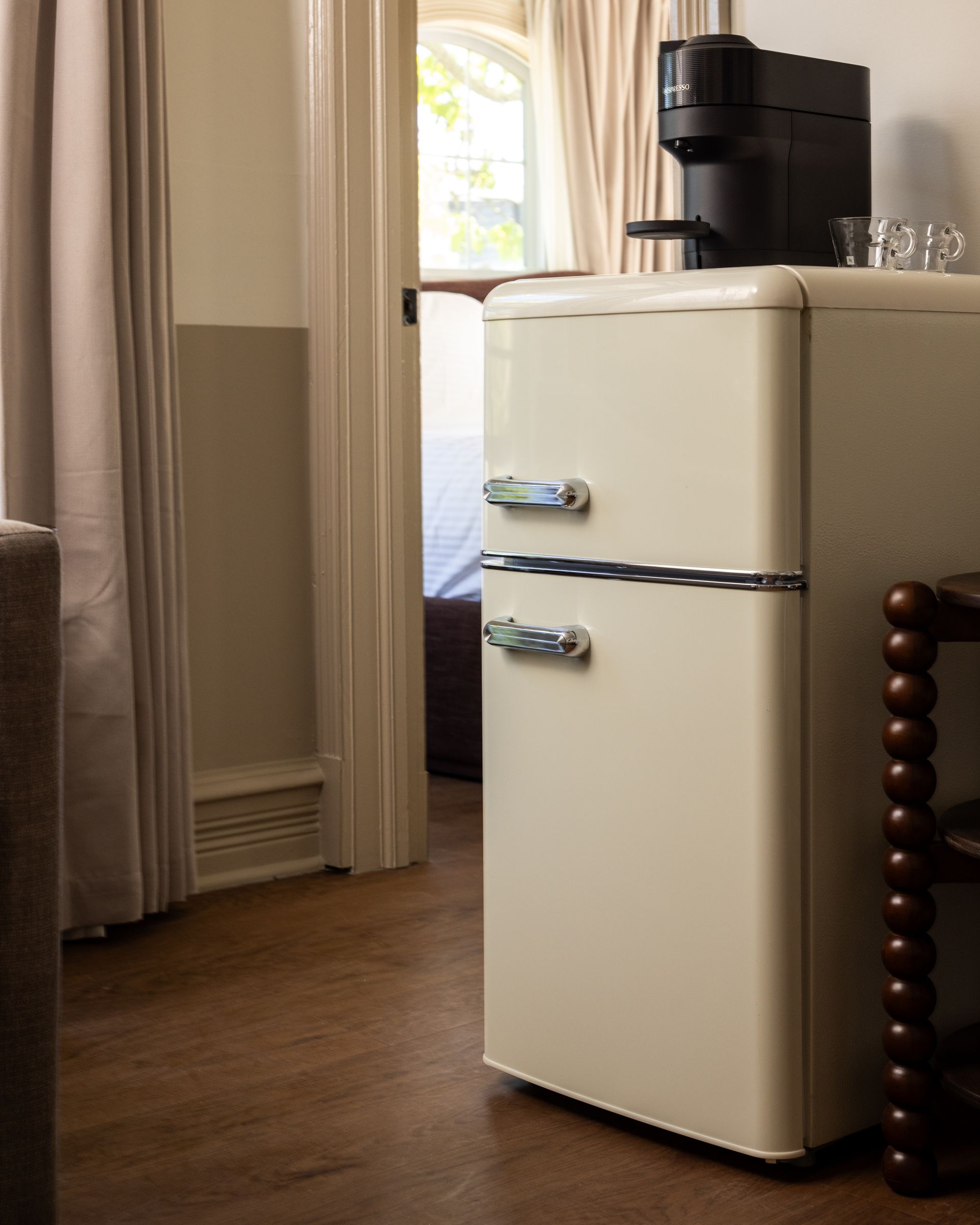 Cream retro refrigerator in a cozy room beside curtains and a window, with a black appliance on top.