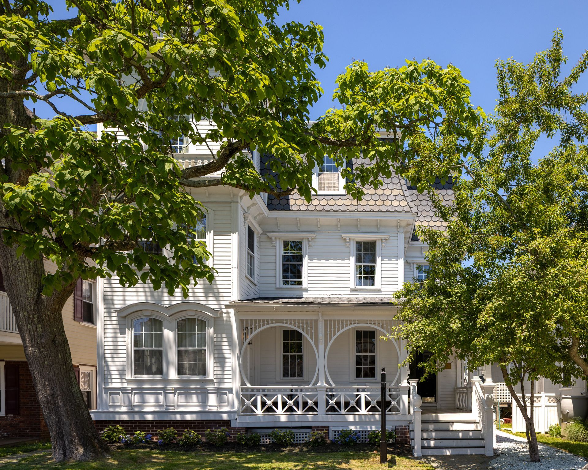White two-story Victorian house with a porch, framed by leafy green trees on a sunny day