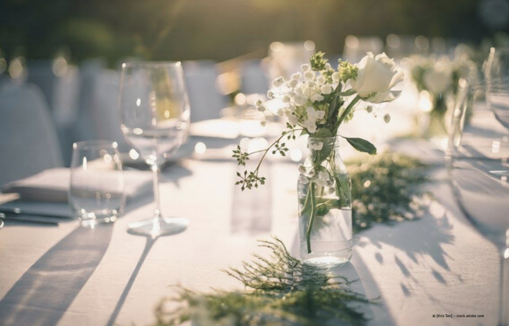 Tischdekoration für eine Hochzeit mit weißen Blumen in einem Glasgefäß, Sonnenlicht und Gedecken.