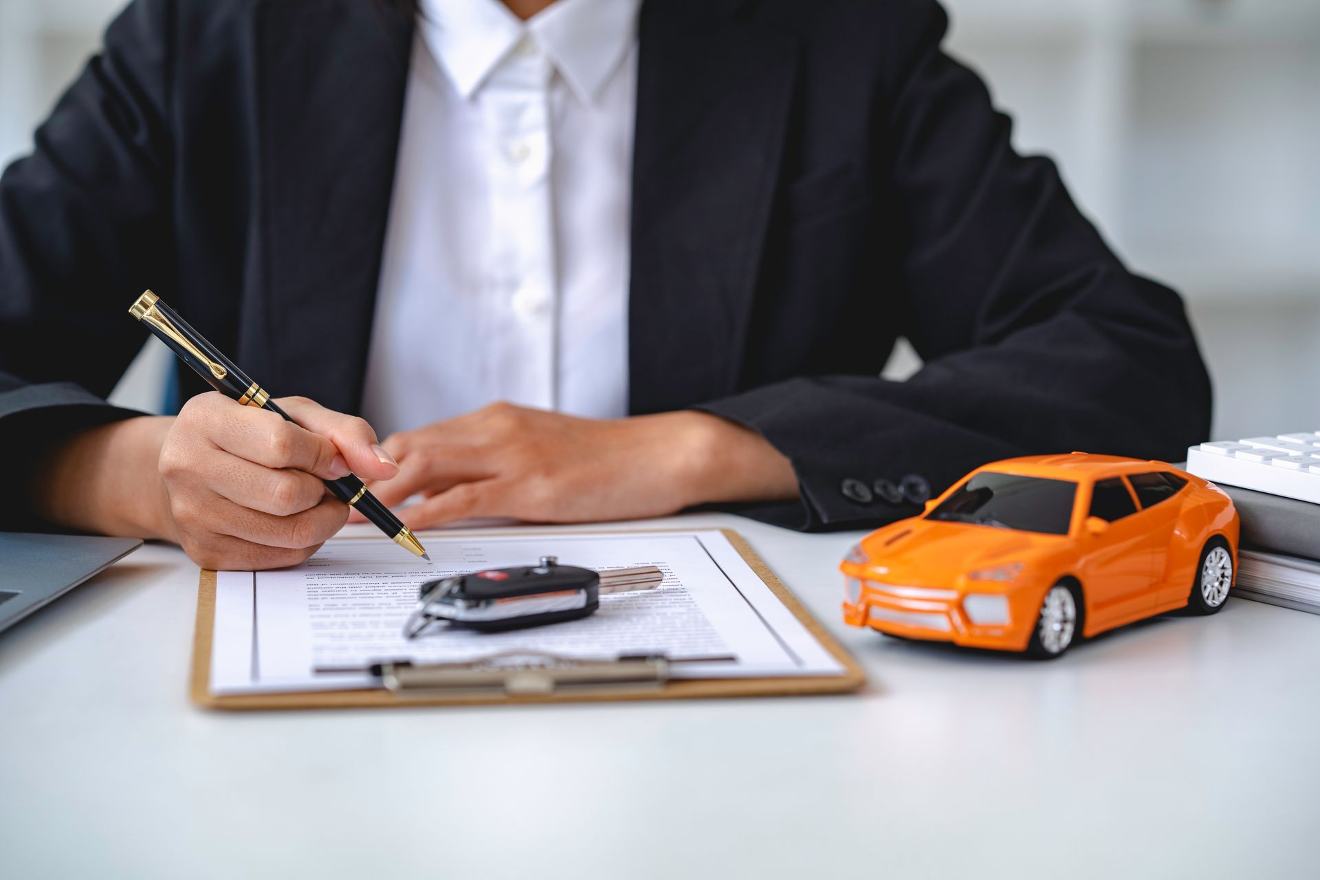 Une personne en costume signe des papiers, des clés de voiture et une petite voiture orange sont posées sur un bureau.