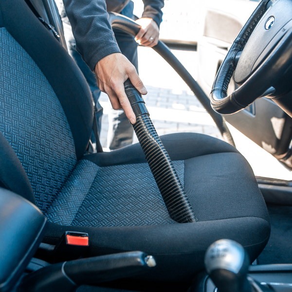 Personne passant l'aspirateur à l'intérieur d'une voiture ; la buse est proche du siège en tissu noir.
