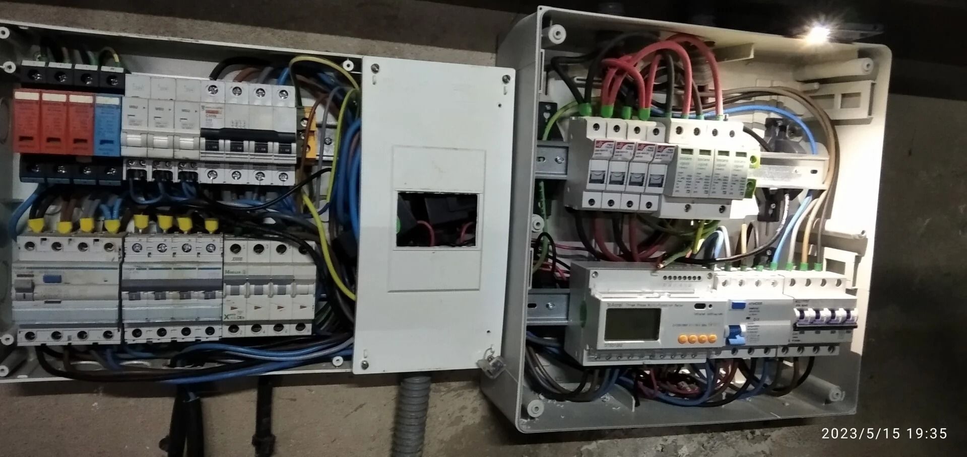 Paneles de control eléctrico abiertos con interruptores, cableado y un medidor central en una pared.