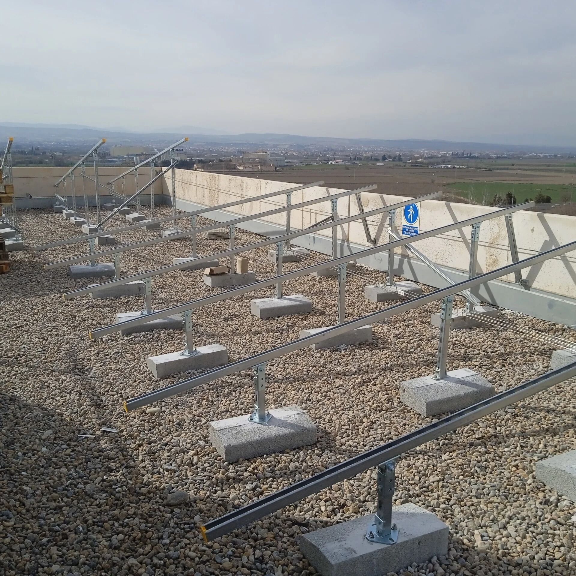 Estructuras de montaje de paneles solares en un tejado de grava con un paisaje lejano bajo un cielo brumoso.