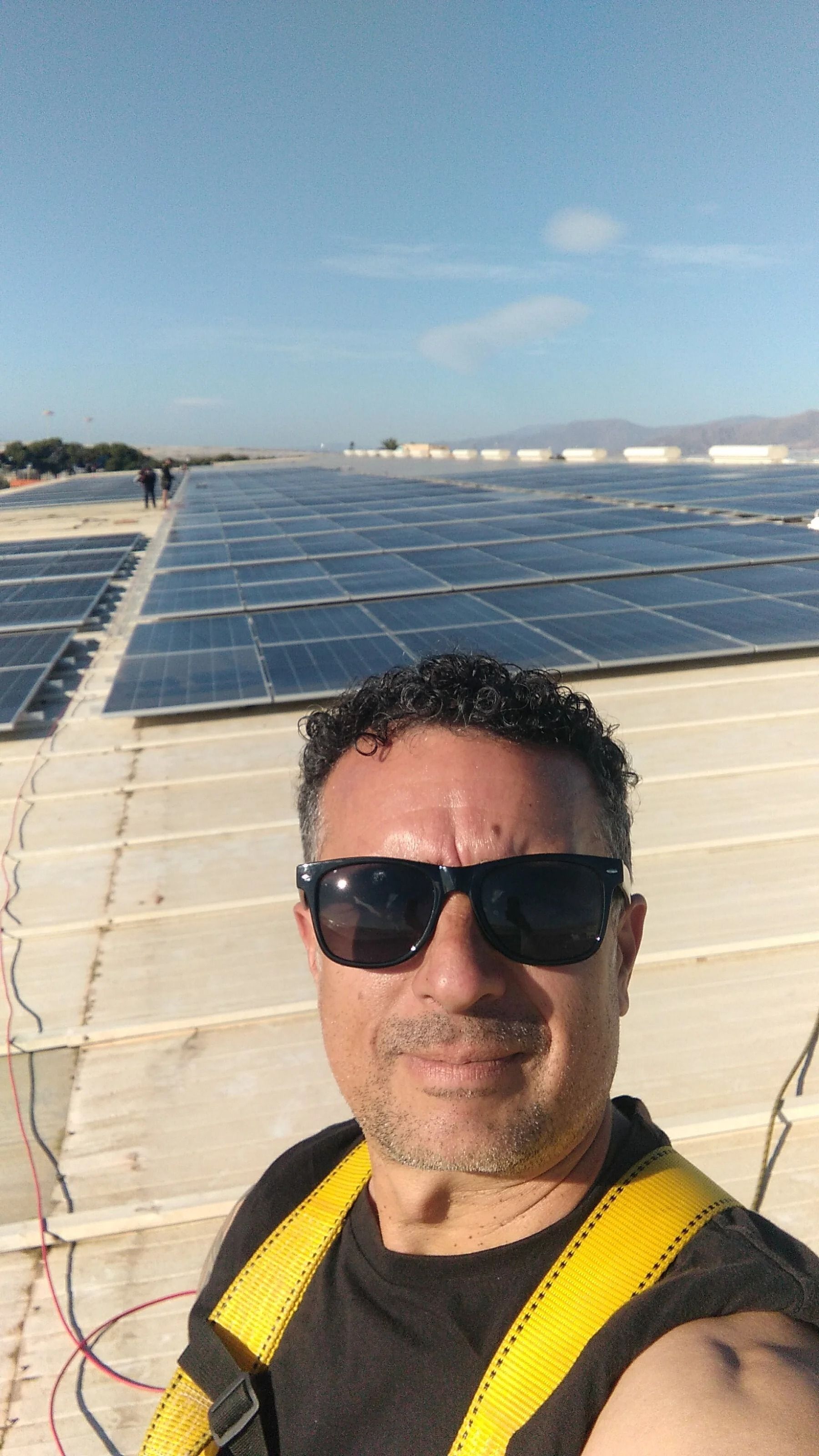 Un hombre se toma una selfie en una azotea con paneles solares bajo un cielo azul despejado.