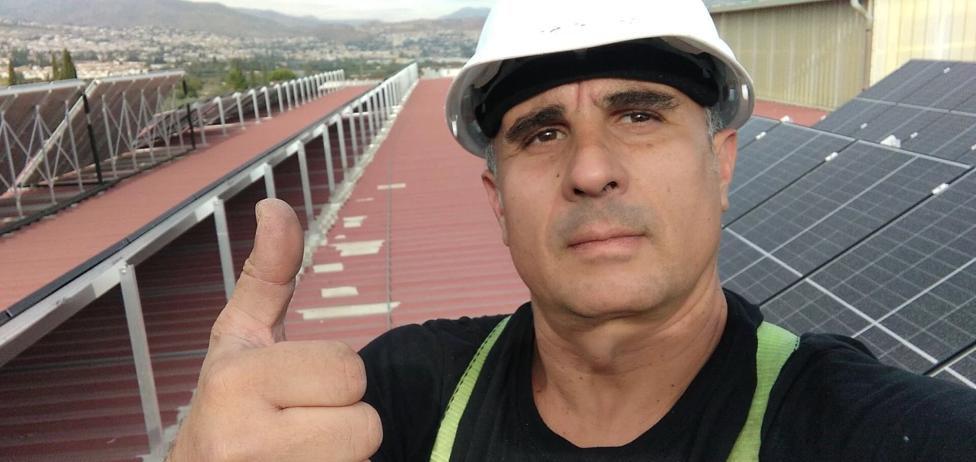 Hombre con casco levanta el pulgar junto a paneles solares en la azotea de un edificio.
