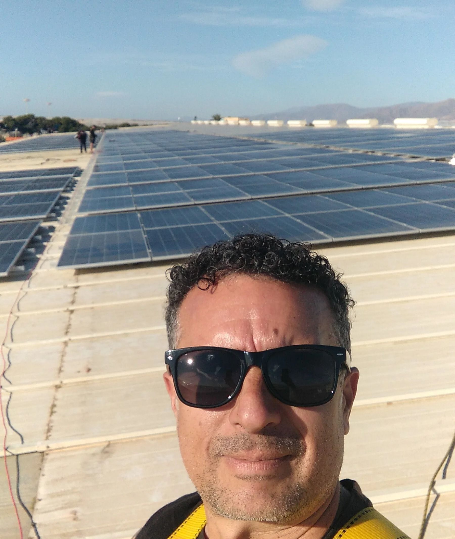 Selfie con gafas de sol de fondo, con hileras de paneles solares que se extienden por un luminoso espacio al aire libre.