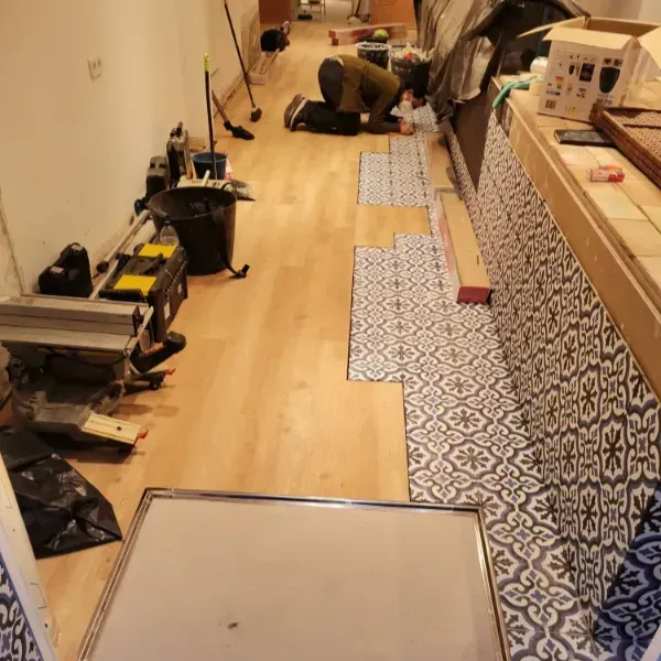 Una persona se arrodilla sobre un piso de madera clara, instalando baldosas estampadas a lo largo de un mueble de cocina