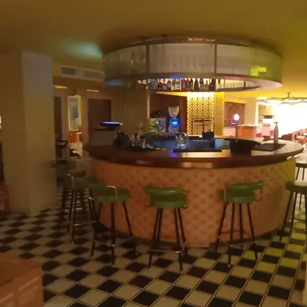 Una barra circular de madera con taburetes verdes y un estante colgante, colocada sobre un suelo a cuadros blancos y negros.