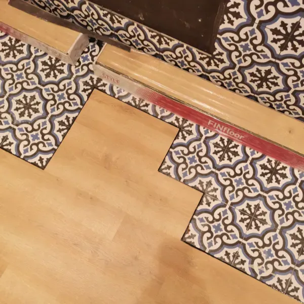 Una tira de transición separa un piso laminado de un piso de baldosas geométricas estampadas en azul y blanco.