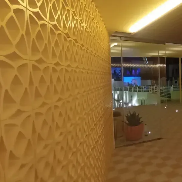 Una mampara geométrica texturizada de color crema junto a una sala de estar interior con paredes de vidrio por la noche.