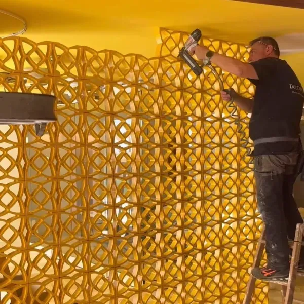 Un trabajador se encuentra de pie sobre una escalera y utiliza una pistola de clavos para fijar paneles de pared geométricos