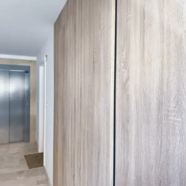 Un pasillo moderno con paredes revestidas de madera clara en primer plano y una puerta de ascensor de metal.