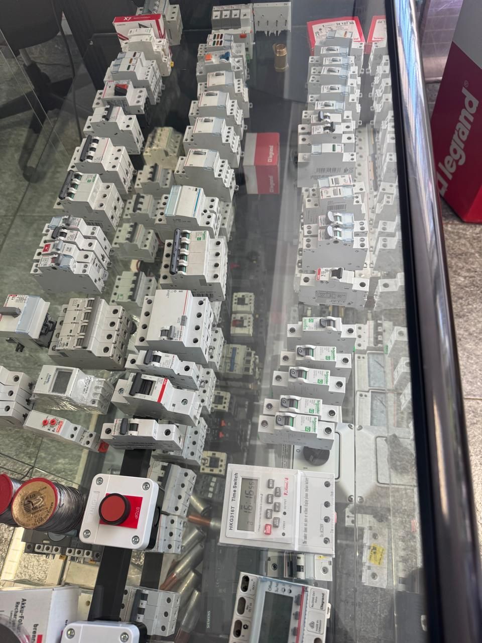 Exposição de componentes elétricos: disjuntores, contatores e temporizadores brancos atrás de um vidro.