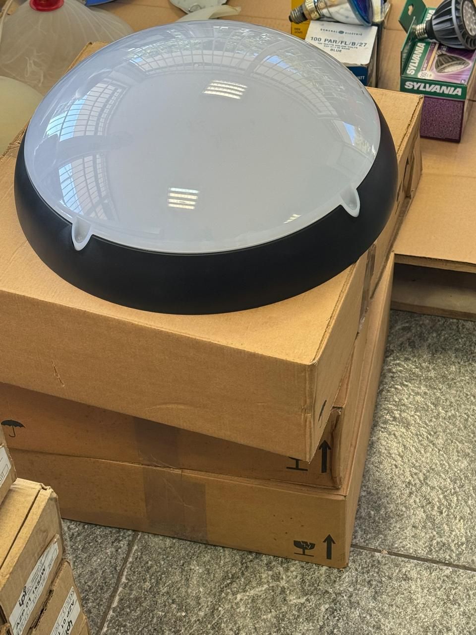Luminária em forma de cúpula com borda preta repousa sobre duas caixas de papelão, com outras caixas e objetos ao fundo.