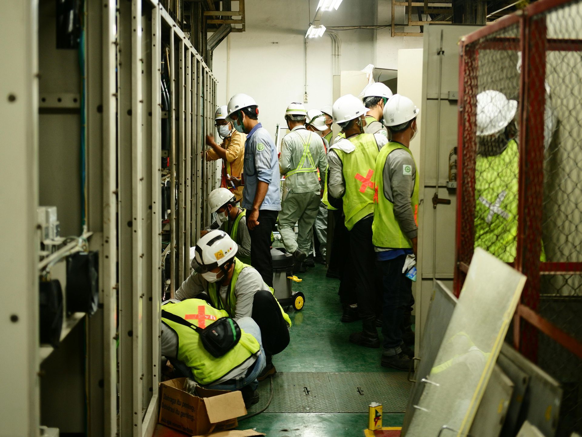 Operários com capacetes e coletes de segurança inspecionam equipamentos elétricos em um espaço industrial estreito.