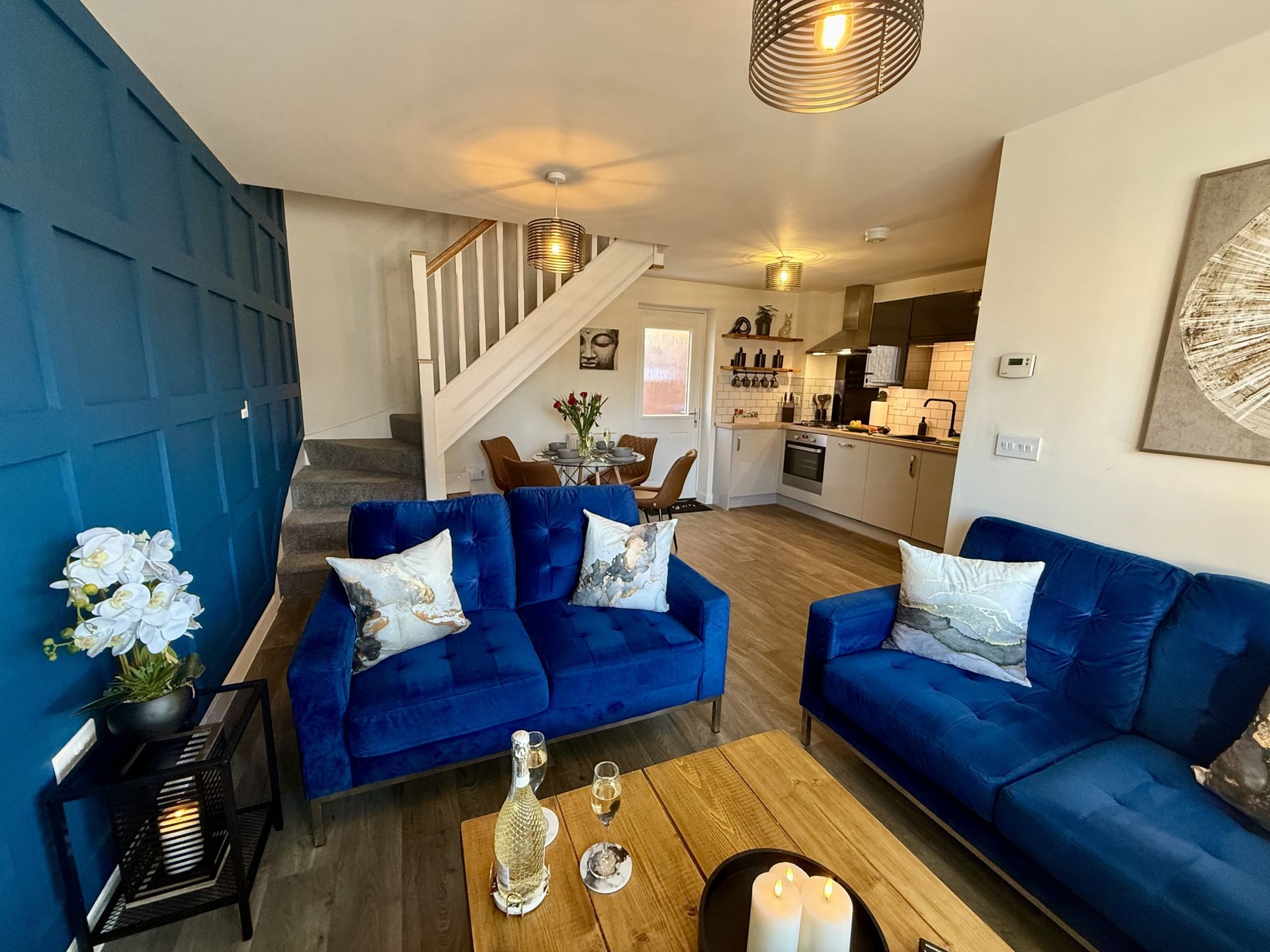 A bright living room with deep blue walls, two matching blue velvet sofas, a wooden coffee table, and an open kitchen.