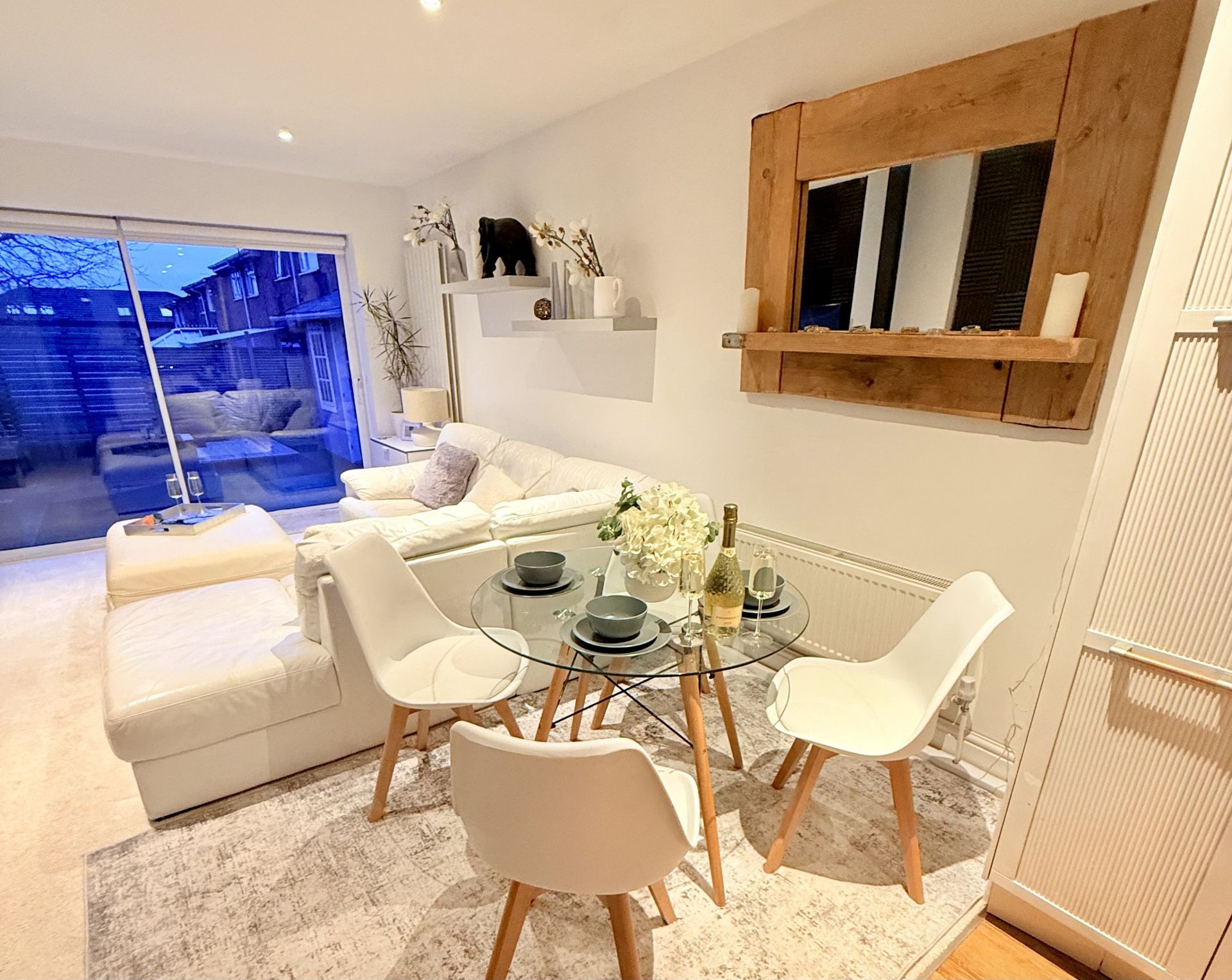A small dining area with a round glass table, three white chairs, and a rustic wooden mirror on a white living room wall.