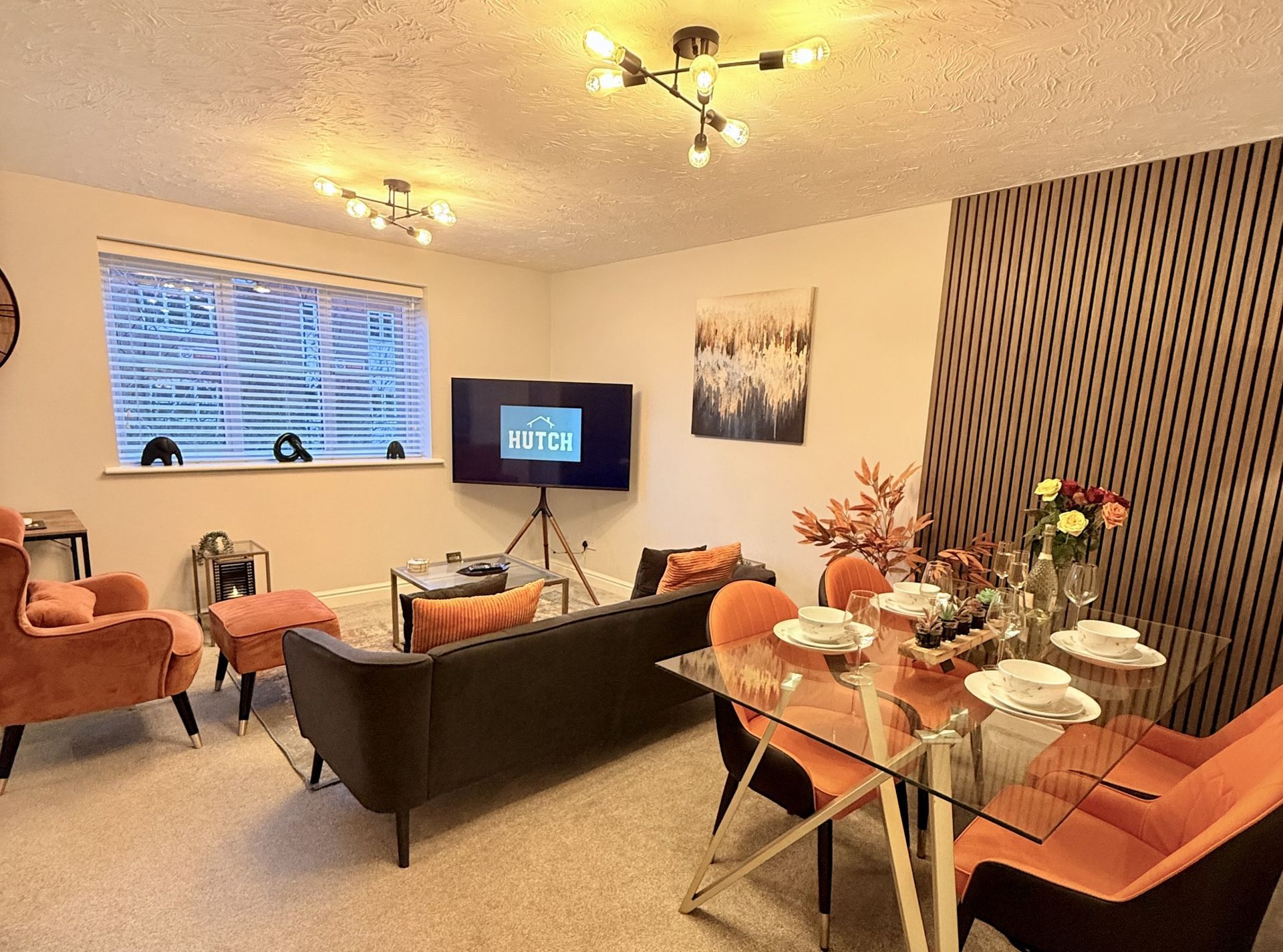 Living room and dining area with orange chairs, a dark sofa, a television on a stand, and modern ceiling lighting.