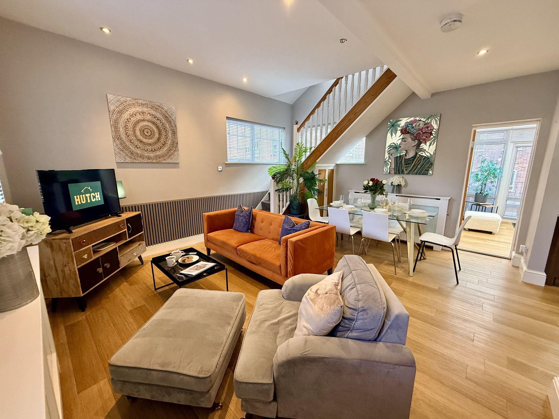 A bright, open-plan living room with wood floors, an orange sofa, grey armchair, television, and a dining area.