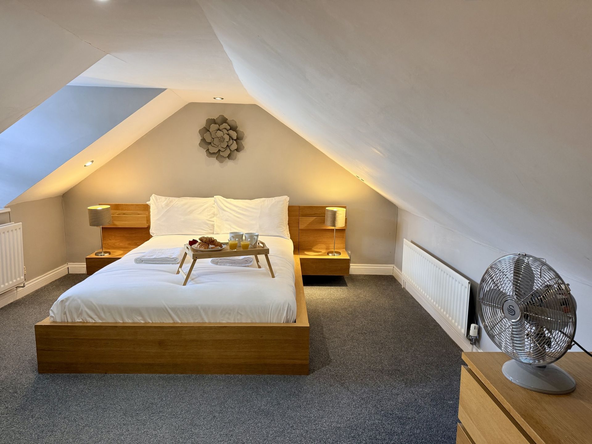 An attic bedroom with a light wooden bed frame, white bedding, grey carpet, a desk fan, and soft ambient lighting.