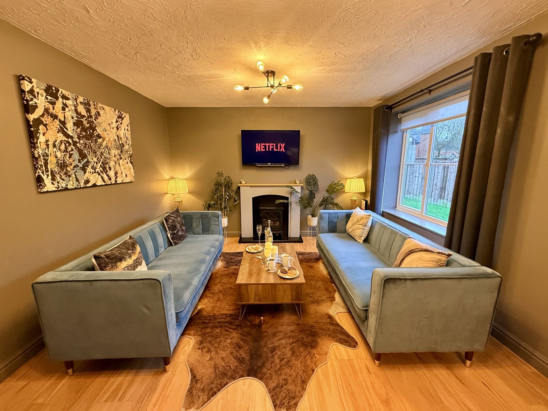 A living room features two teal sofas facing a wooden coffee table, a fireplace with a TV, and a brown cowhide rug.