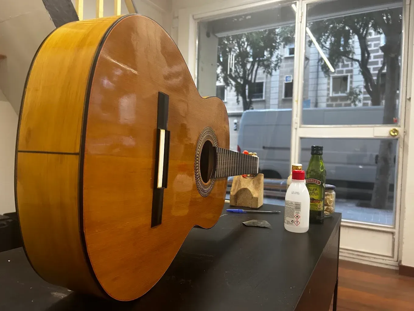 Una guitarra acústica descansa sobre una mesa oscura frente a una ventana, con una botella y un pequeño recipiente a su lado.