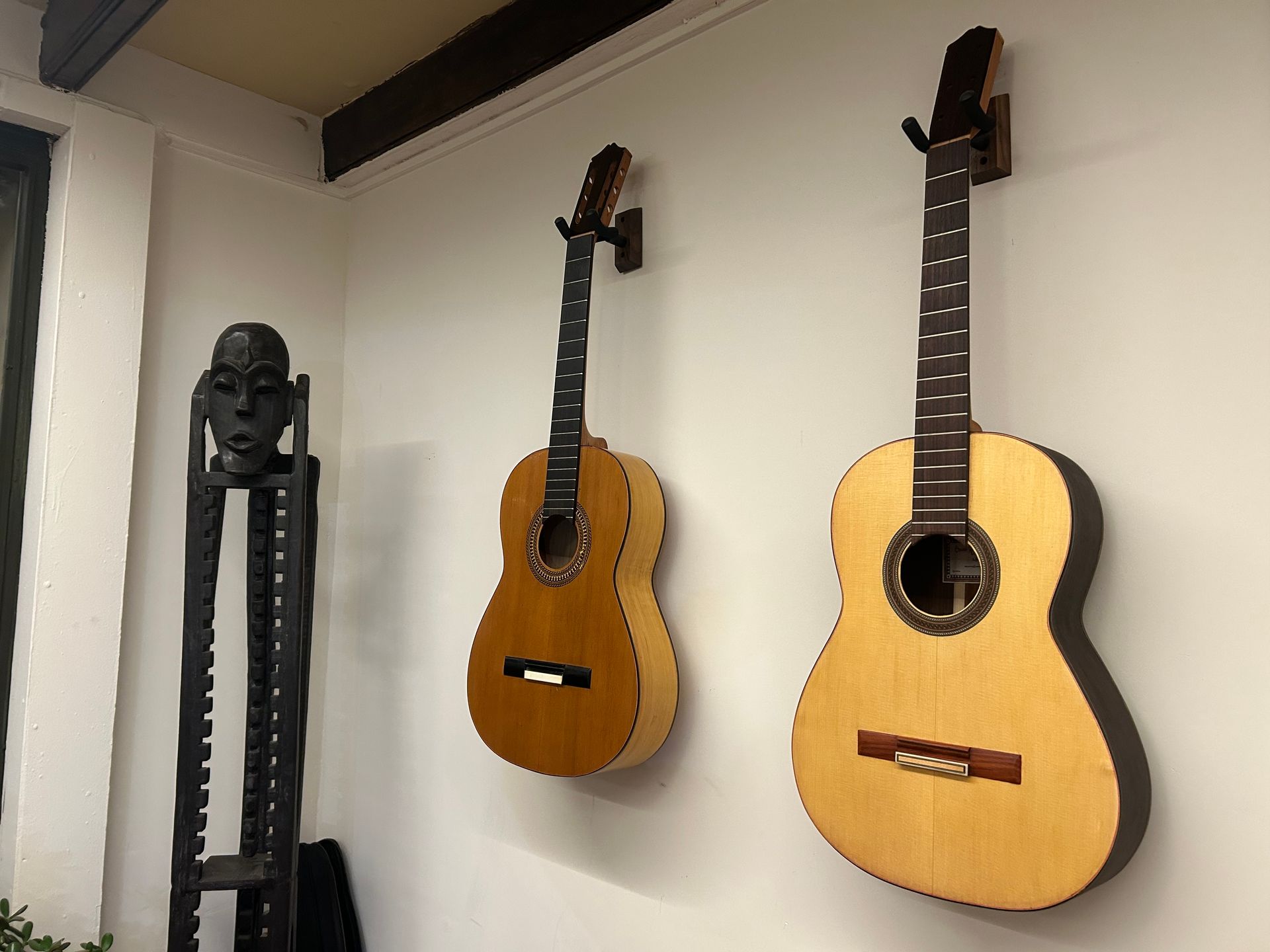 Dos guitarras acústicas cuelgan de una pared blanca junto a una alta escultura de madera oscura con forma de tótem.