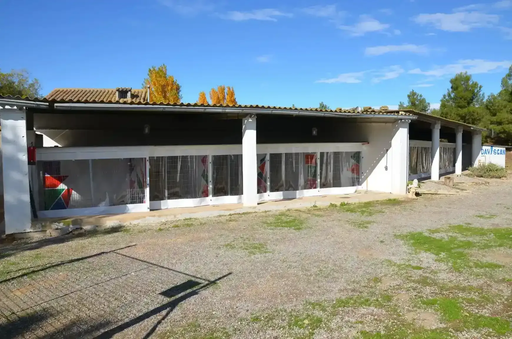 Edificio largo y blanco con varias ventanas transparentes bajo un cielo azul; grava en primer plano.