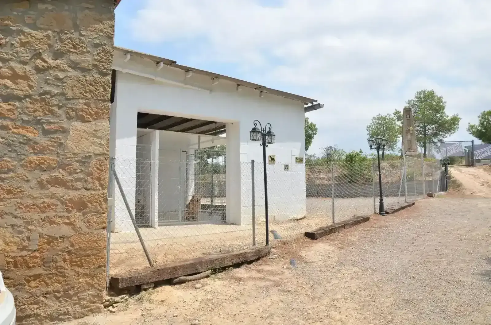 Edificio blanco con puerta abierta, cerca de alambre, camino de grava y muro de piedra en un día soleado.