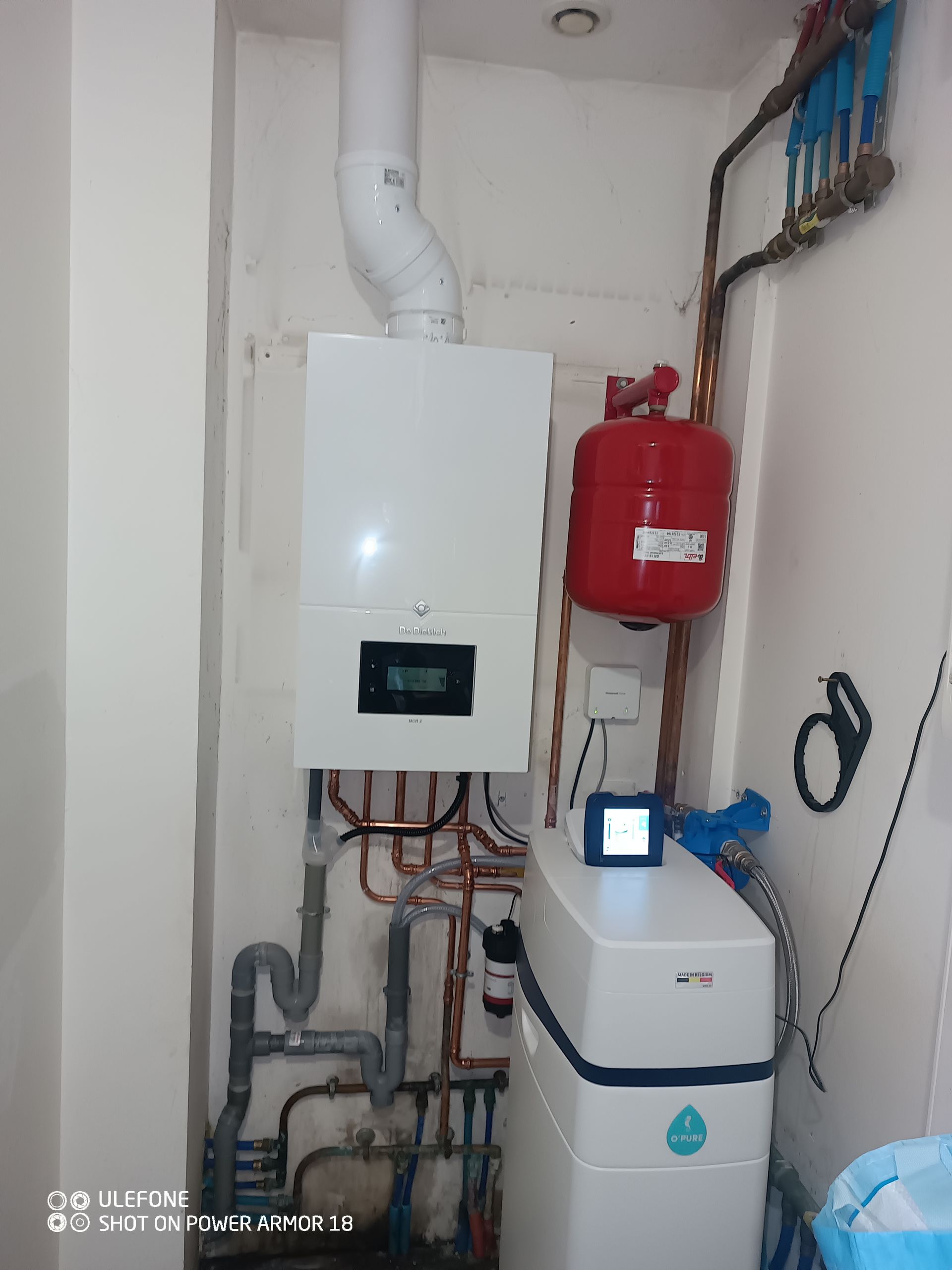 Une chaudière murale blanche, un vase d'expansion rouge et un adoucisseur d'eau installés dans un placard technique.