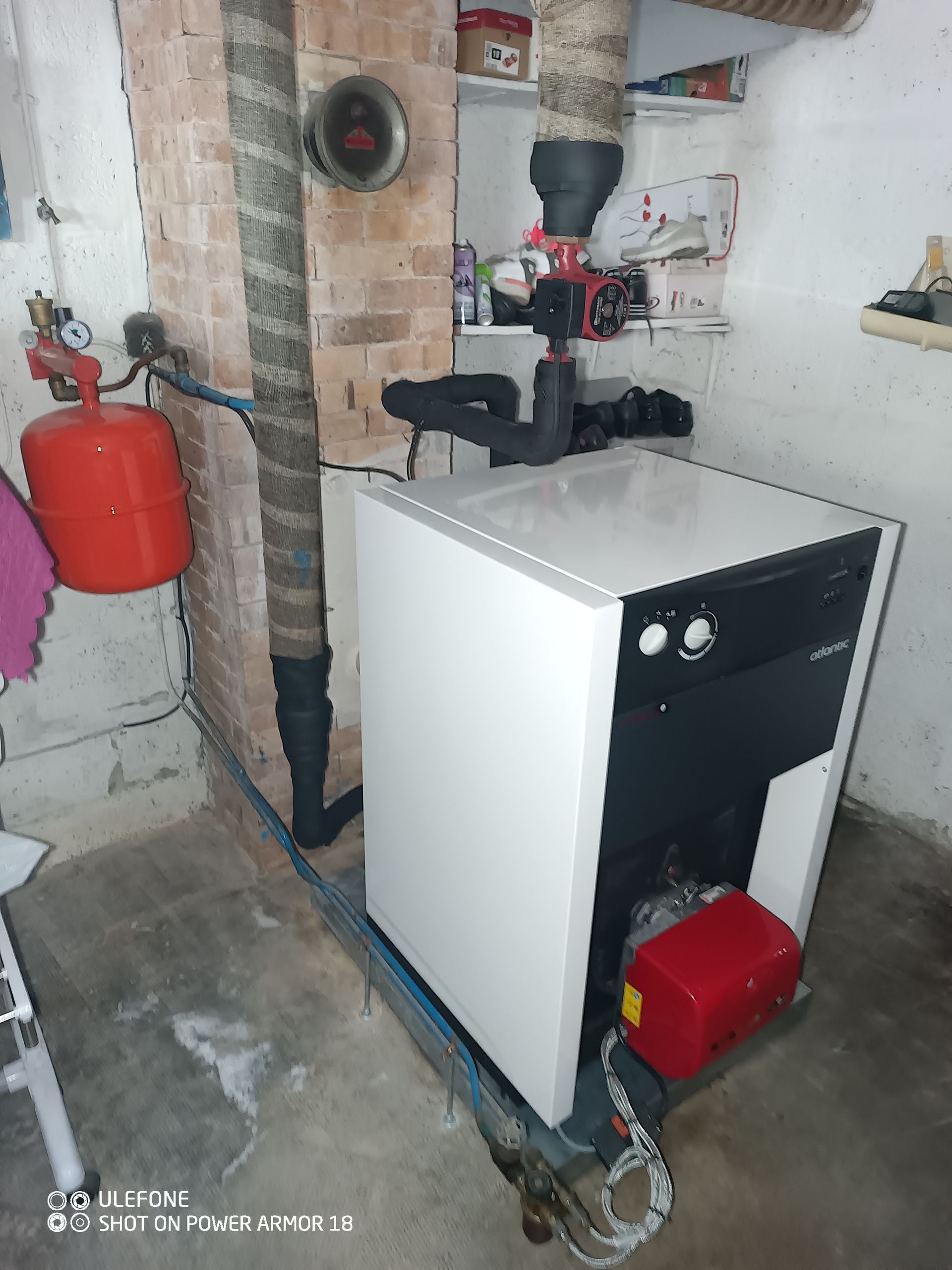 Une chaudière blanche sur pied dans un sous-sol, avec un vase d'expansion rouge à gauche et une tuyauterie isolée au-dessus.