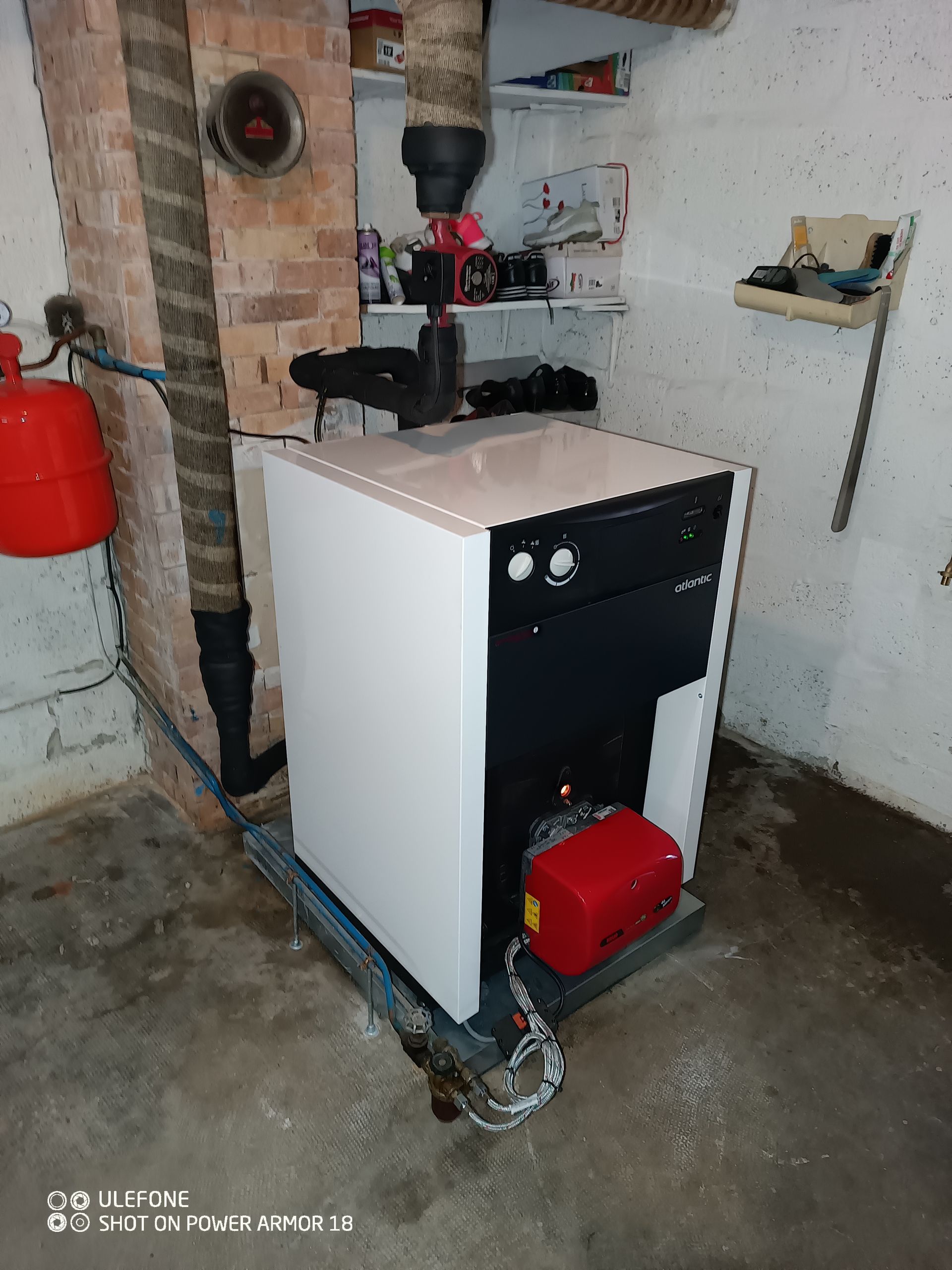 Une chaudière blanche et noire sur pied, équipée d'une pompe à combustible rouge, se trouve dans un sous-sol près d'une cheminée en briques et d'un réservoir rouge.