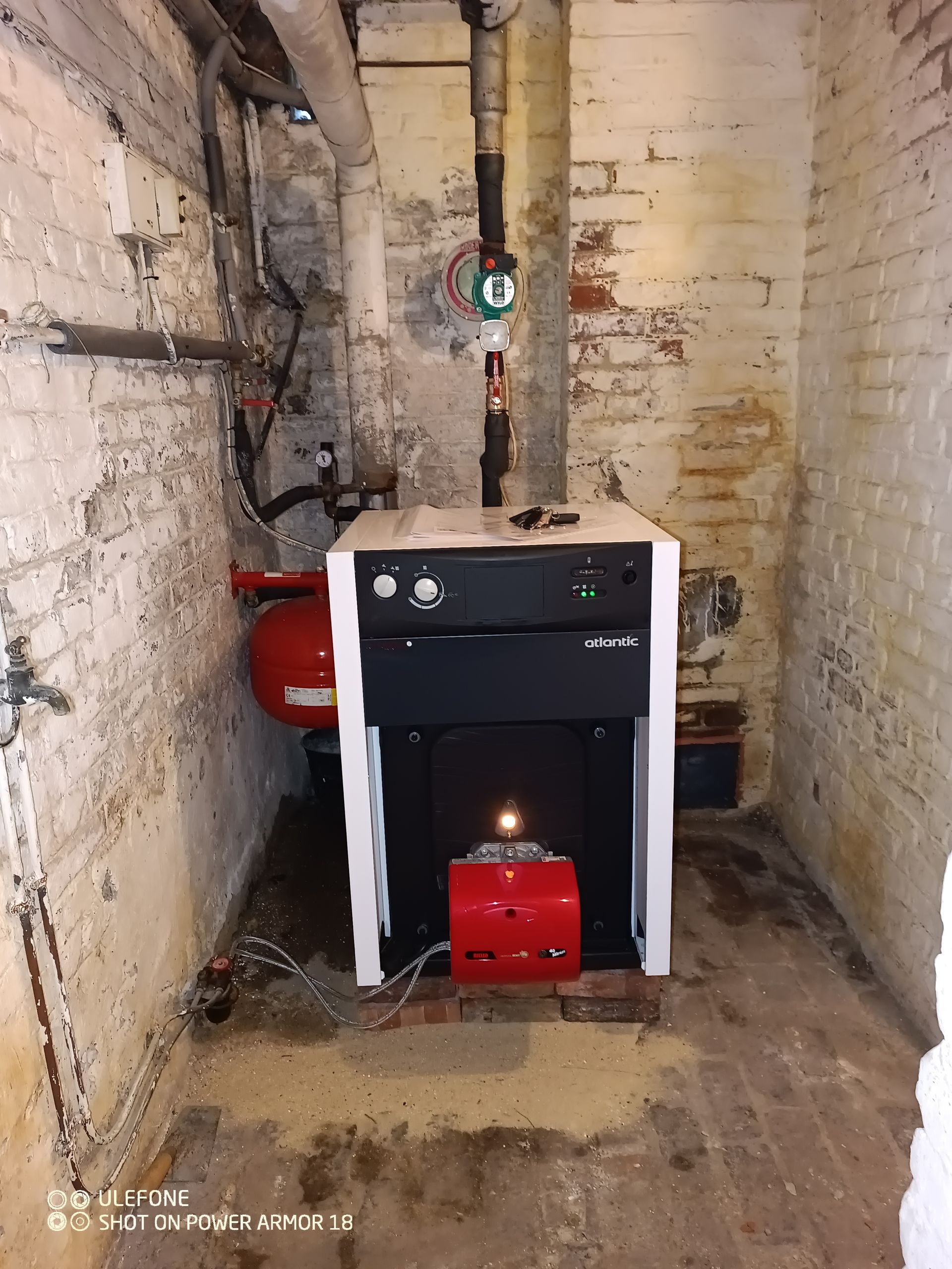 Une chaudière à fioul noire et blanche avec un brûleur rouge trône dans un sous-sol aux murs de briques texturées et à la tuyauterie apparente.