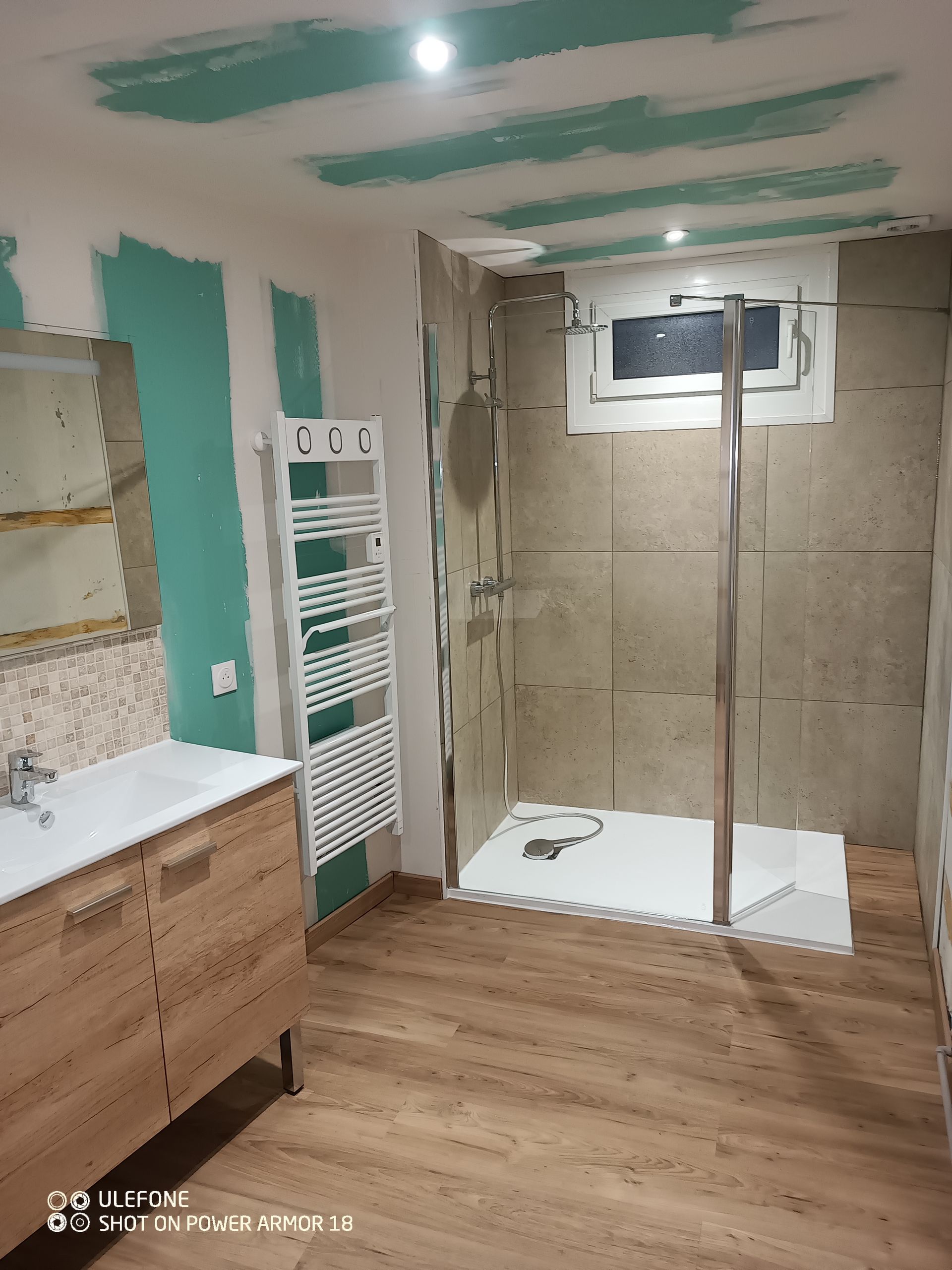 Une salle de bains en cours de rénovation avec un meuble-lavabo en bois, une douche à l'italienne, un sèche-serviettes et des murs partiellement peints en vert.