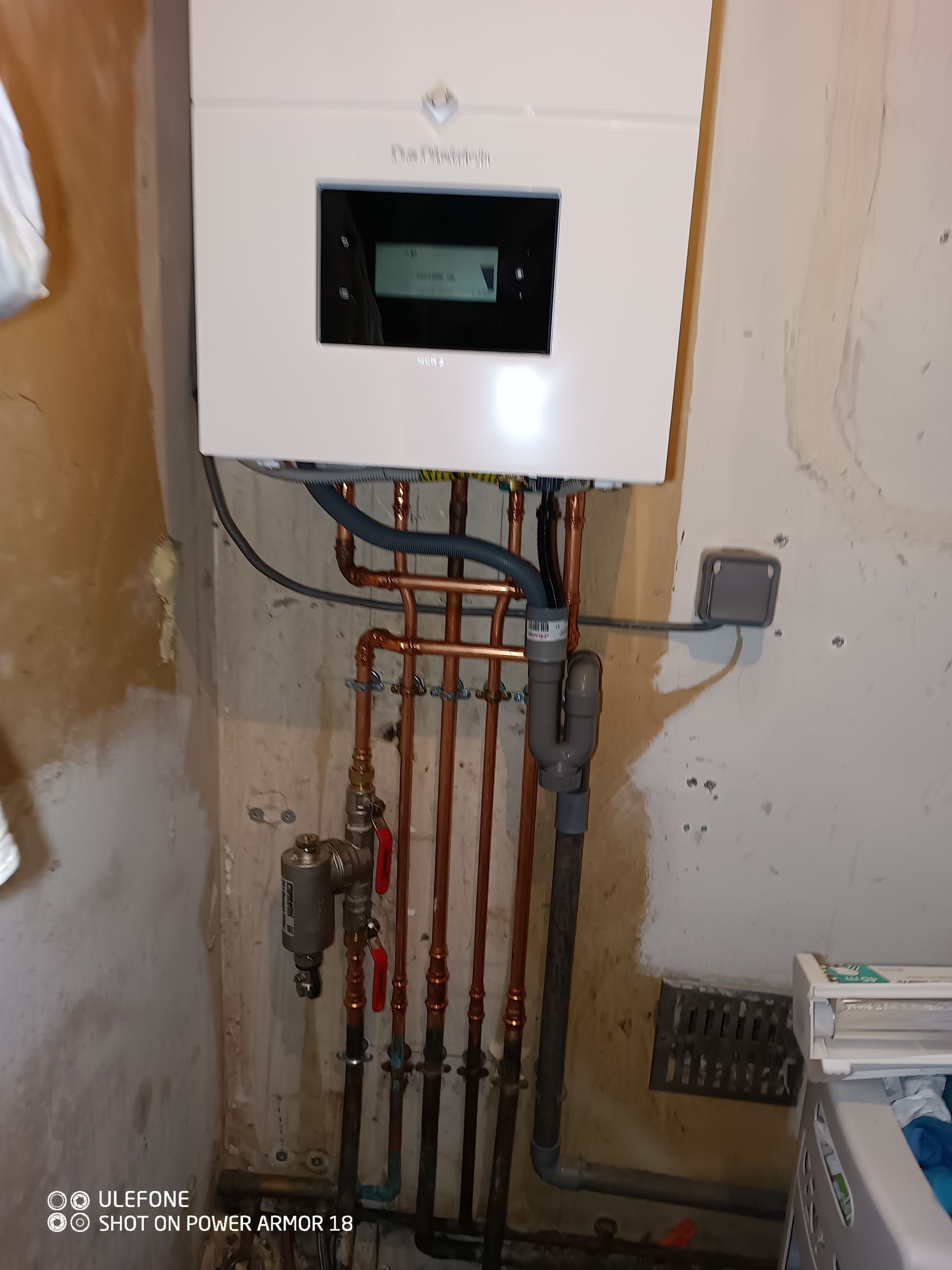 Une chaudière murale blanche pour le chauffage central, avec tuyauterie en cuivre et écran d'affichage, installée sur un mur en plâtre brut.