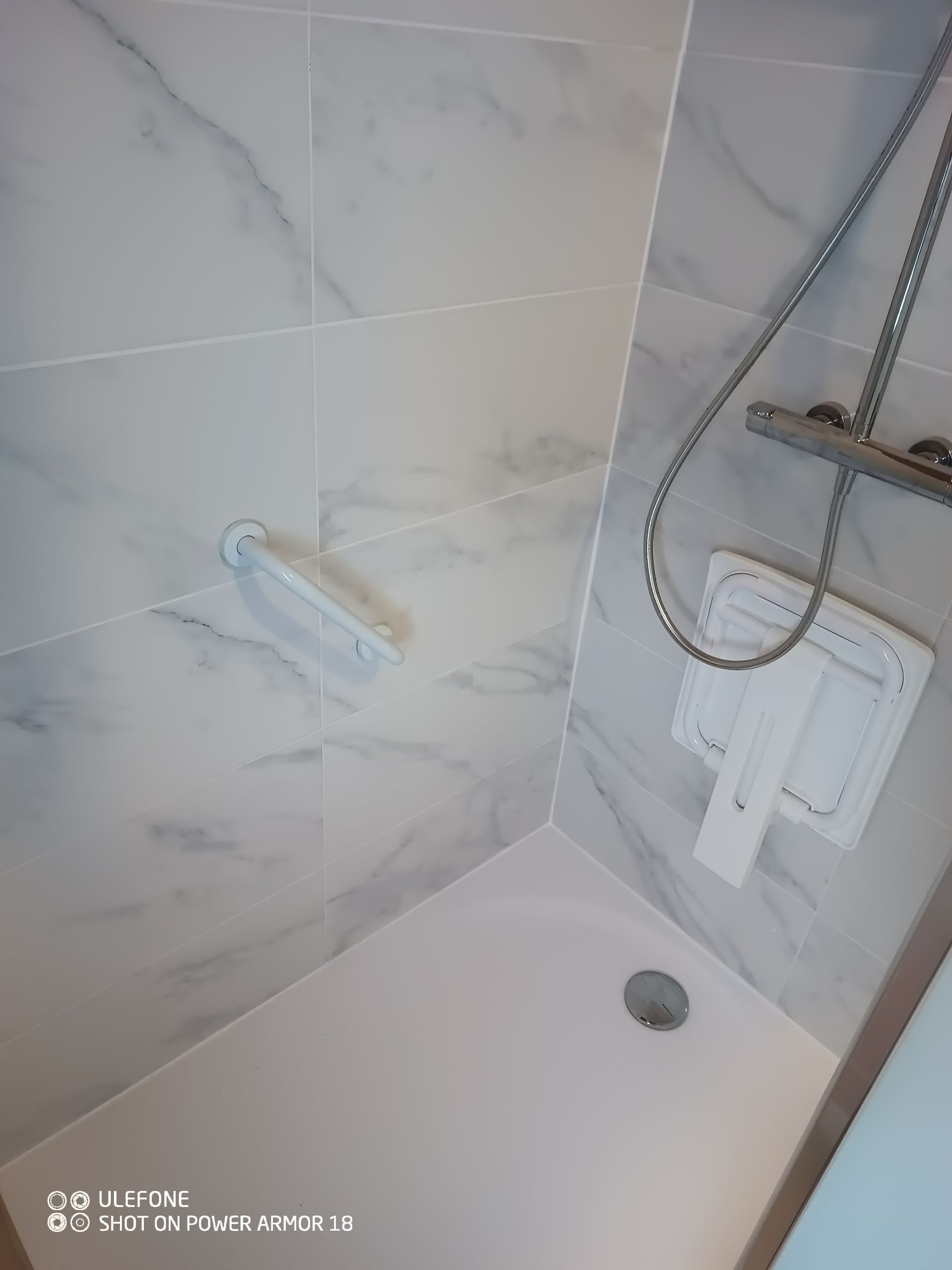 Une cabine de douche avec des carreaux blancs à motif marbre, une barre d'appui d'angle blanche et un siège de douche blanc fixé au mur.