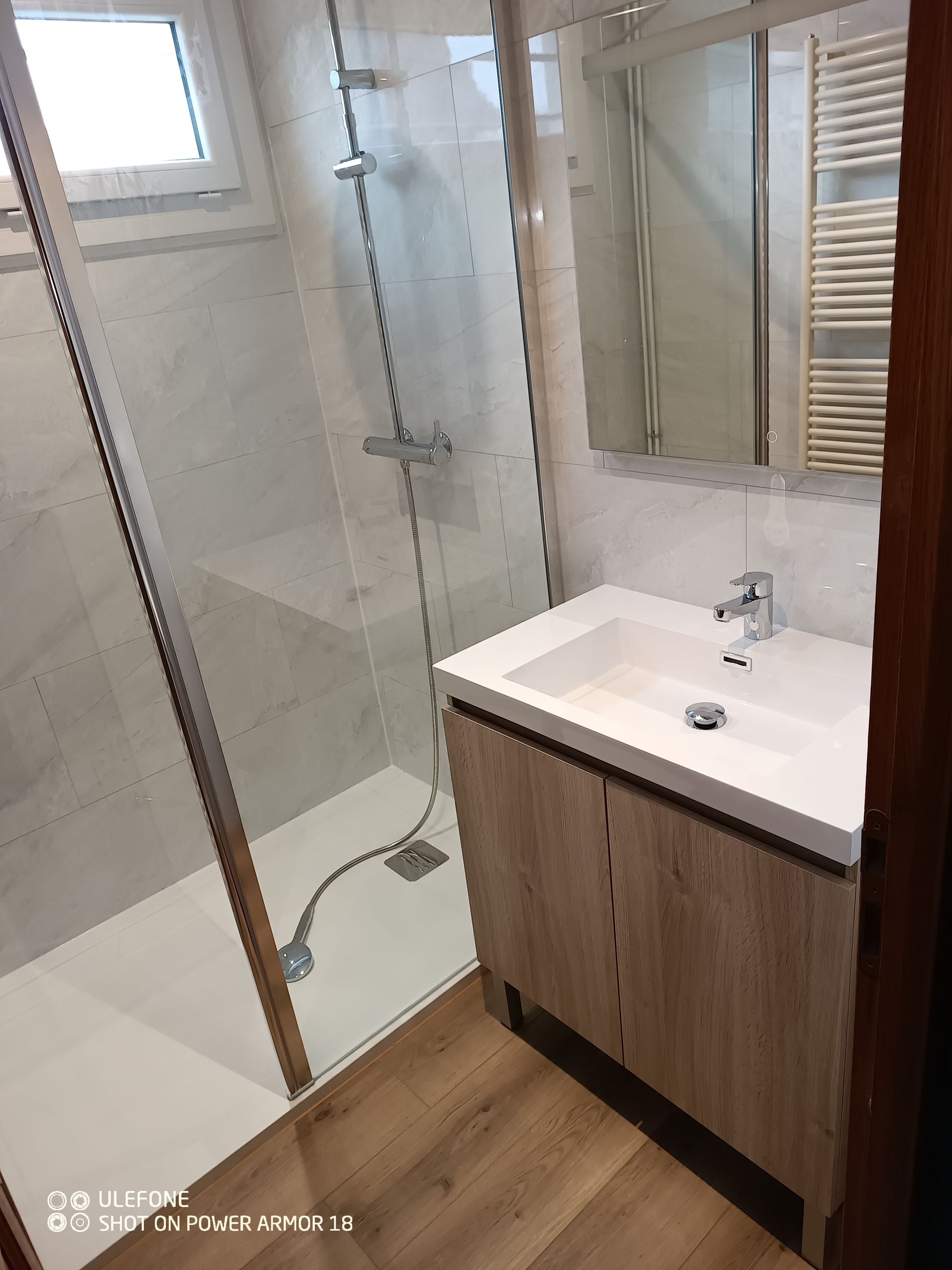 Salle de bains moderne avec douche vitrée, meuble-lavabo en bois clair, lavabo blanc et carrelage clair au sol.