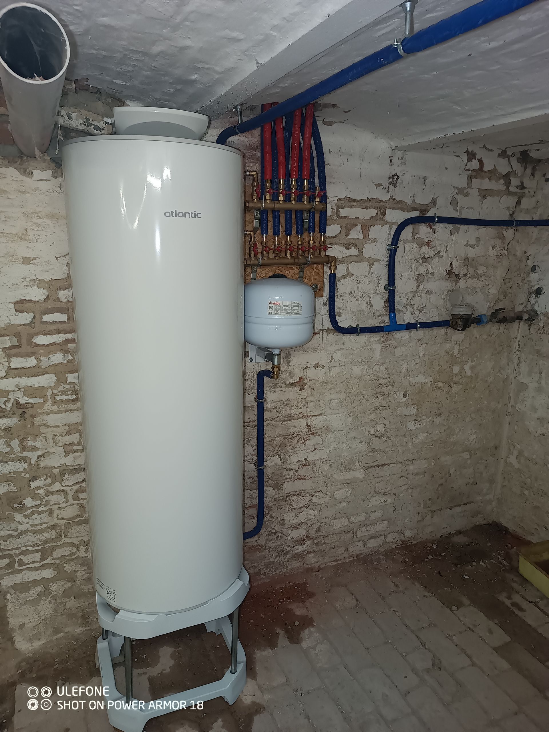 Un réservoir de chauffe-eau blanc se trouve à côté d'un collecteur de tuyaux rouges et bleus fixé au mur et d'un petit vase d'expansion.