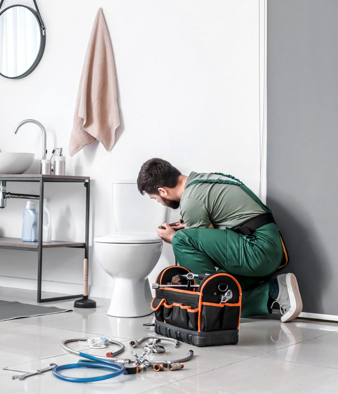 Un plombier en combinaison verte répare des toilettes dans une salle de bains lumineuse, avec une sacoche à outils et du matériel de plomberie éparpillés sur le sol.