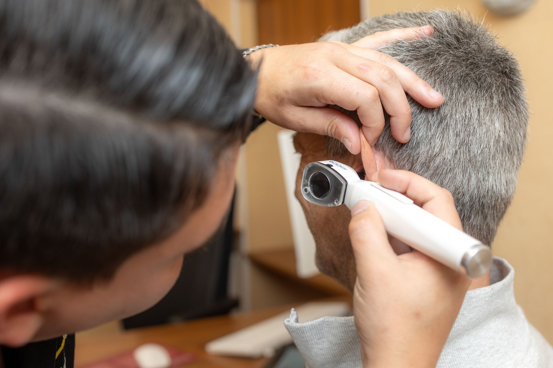 Arzt untersucht das Ohr des Patienten mit einem Otoskop. Sitzender grauhaariger Mann, hellhäutiger Arzt schaut zu.