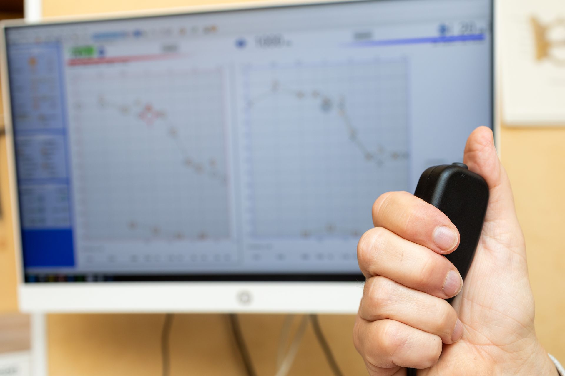 Eine Hand hält eine schwarze Fernbedienung, im Hintergrund ein Computermonitor mit Börsencharts.