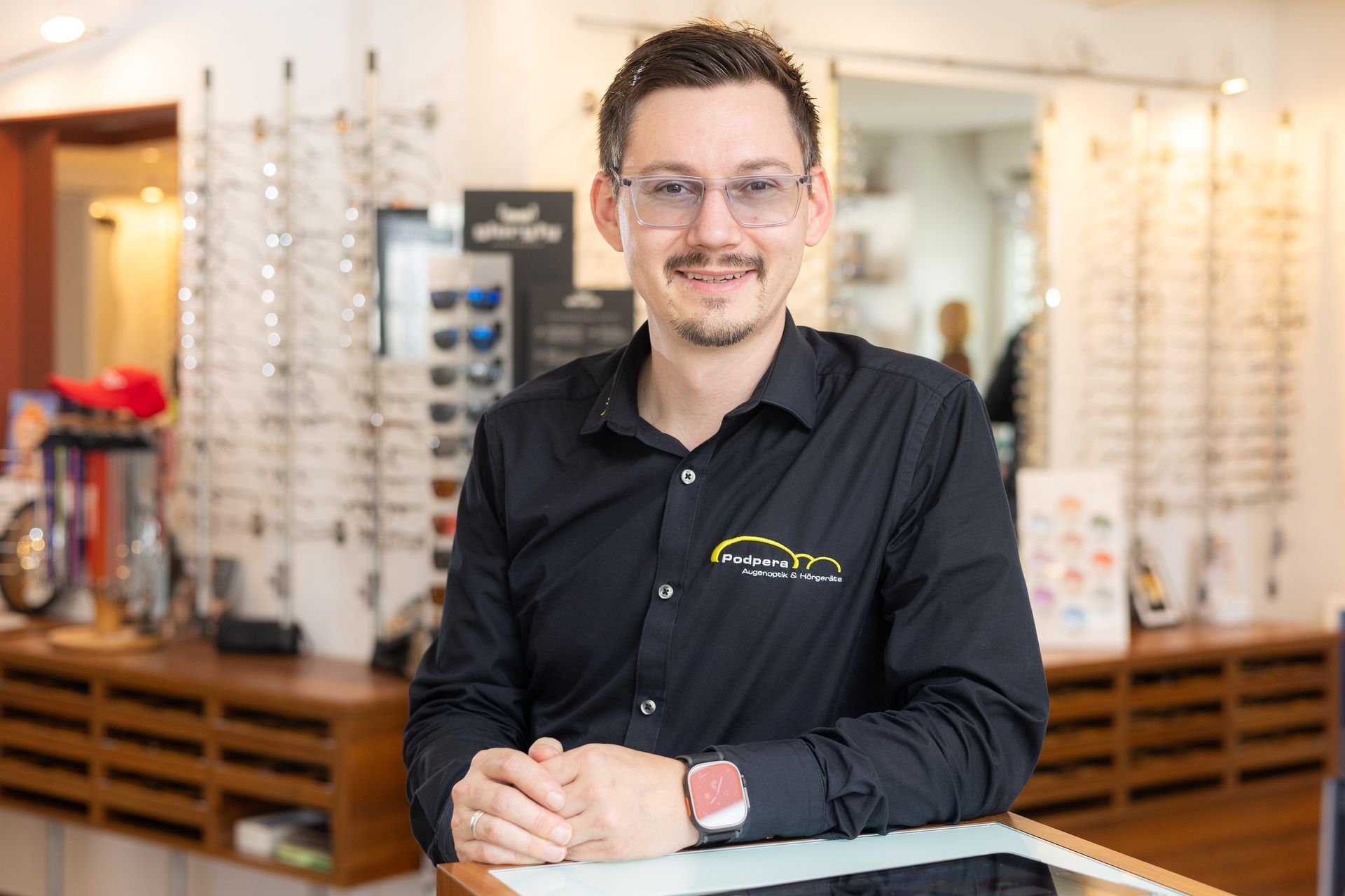 Mann im schwarzen Hemd mit Brille, lächelnd in einem Optikergeschäft.